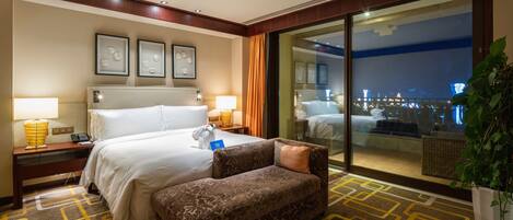 Habitación de hotel con una cama grande, un sofá, un escritorio con lámpara y vista a la ciudad de noche.