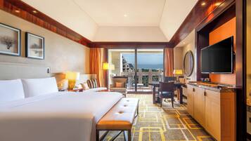 A hotel room with a large bed, a desk with a TV, a balcony with a view, and a patterned carpet.