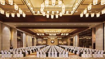 Un amplio salón de conferencias con filas de mesas y sillas, candelabros ornamentales y un gran candelabro.