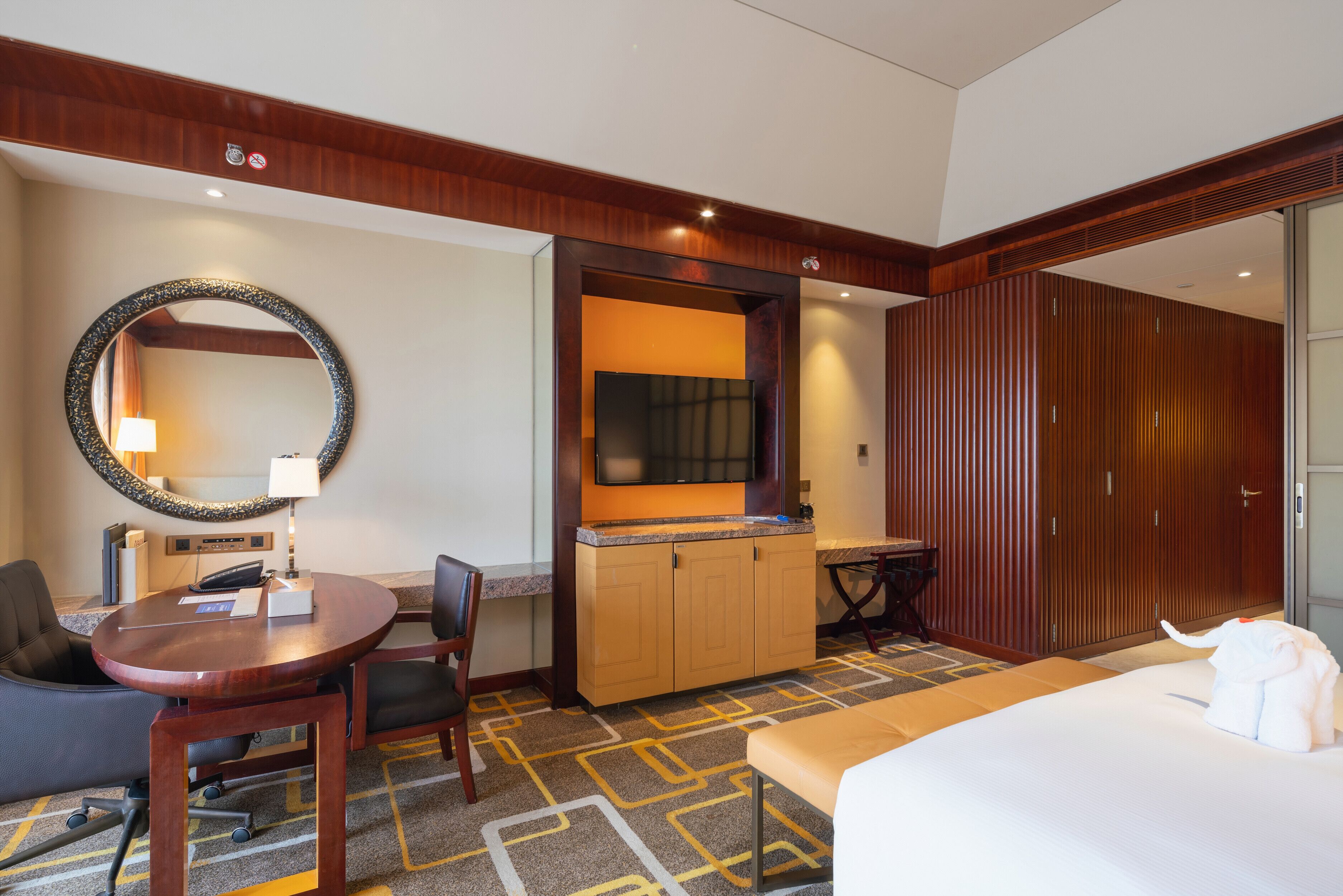 A hotel room with a bed, a desk with chairs, a small kitchenette, a mirror, and a wardrobe.