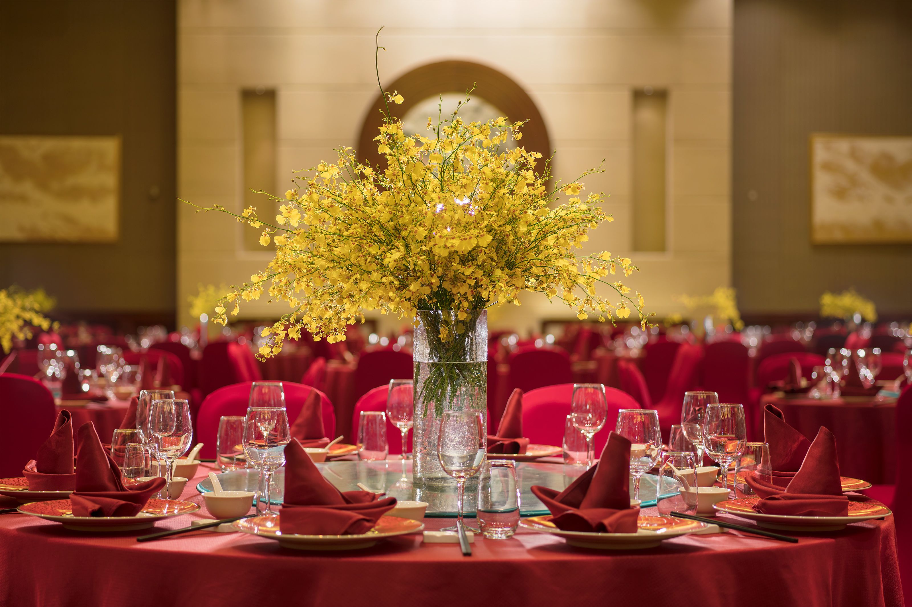 A banquet hall with tables set for a formal event, featuring a central floral arrangement in a tall vase.