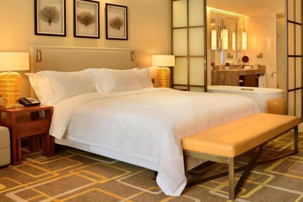 A hotel room with a large bed, bedside tables, a bench, a bathroom with a mirror, and framed artwork on the wall.