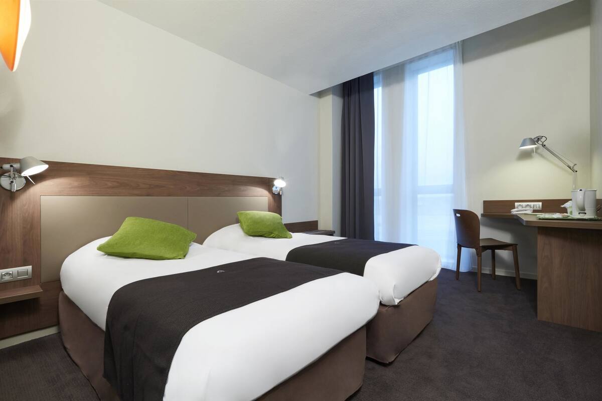 Ein Hotelzimmer mit zwei Betten, einem Schreibtisch, einem Stuhl und einem großen Fenster.