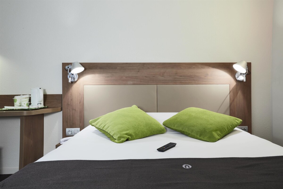 A hotel room with a wooden headboard, two green pillows, a remote control, and a bedside table with a coffee maker and a tissue box.