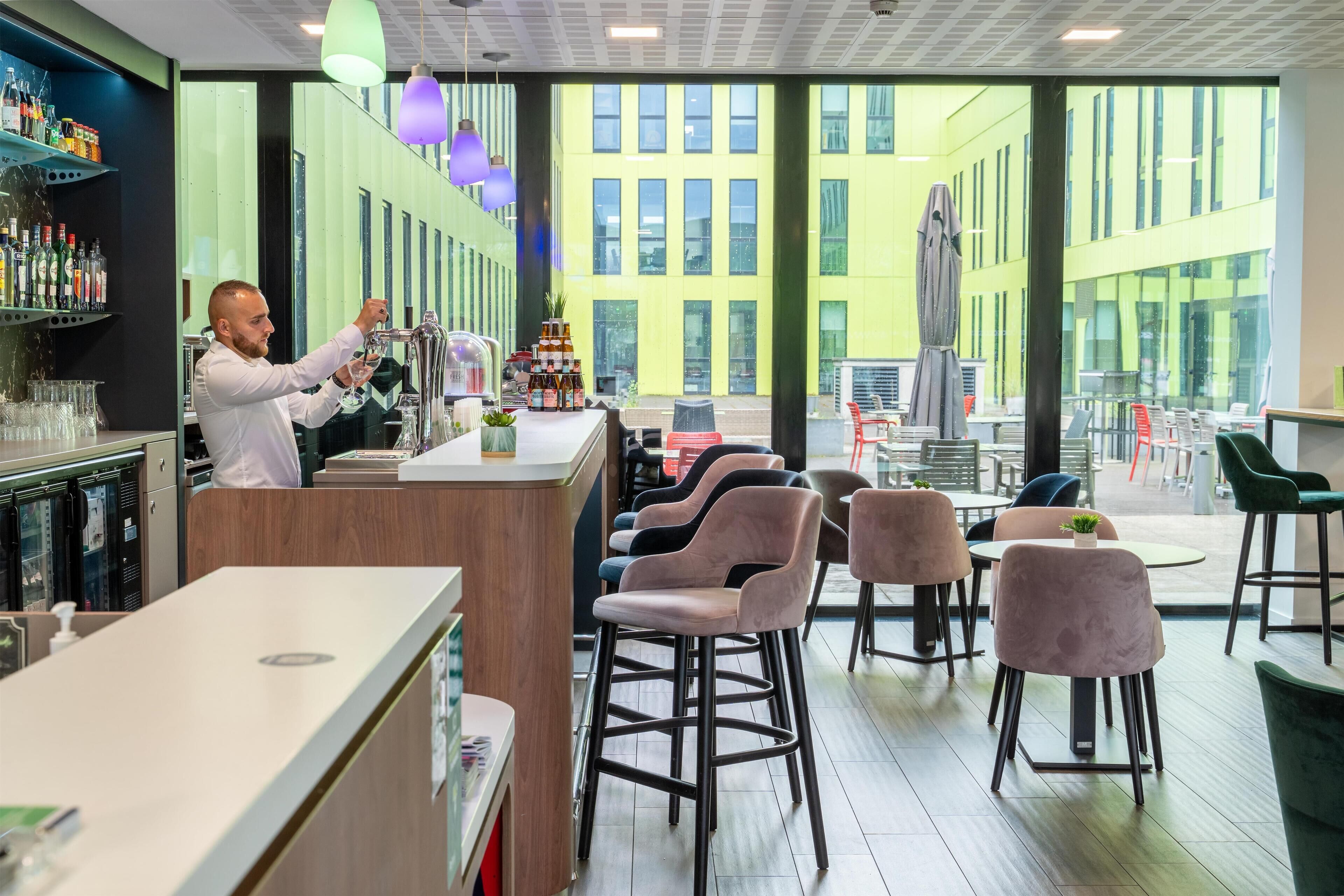 A modern bar area with a bartender preparing drinks, a variety of bottles on shelves, and a seating area with chairs and tables.
