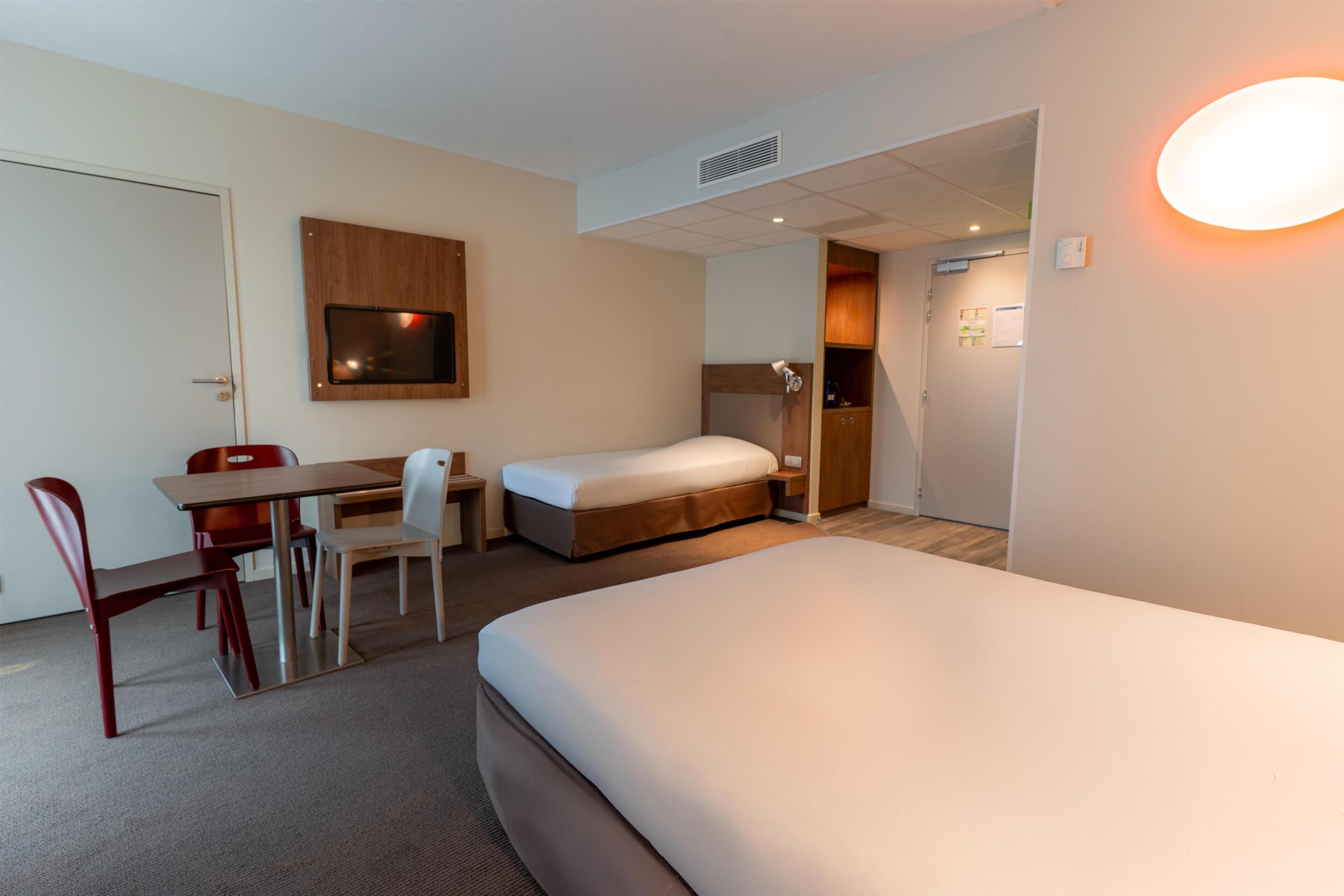 A hotel room with a bed, a desk with a chair, a television, and a bathroom visible through an open door.