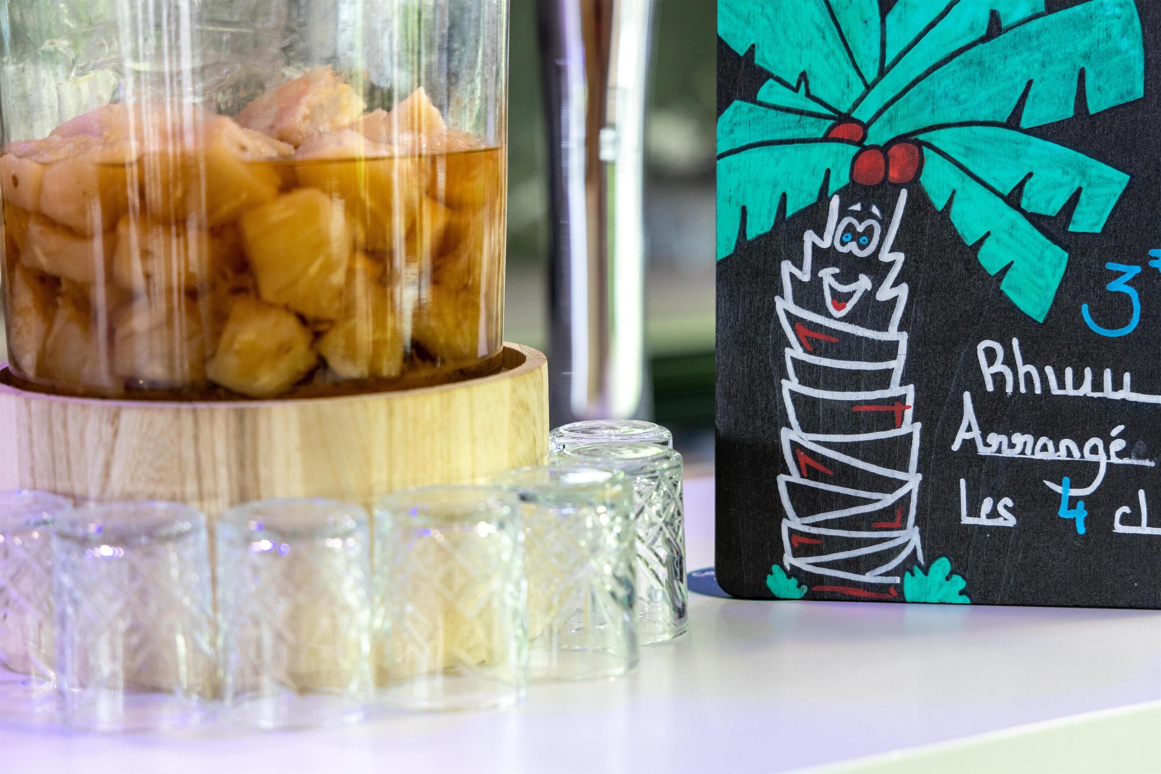 A glass container with a wooden base filled with food items, a blackboard with a drawing of a palm tree and text, and several clear glass cups.