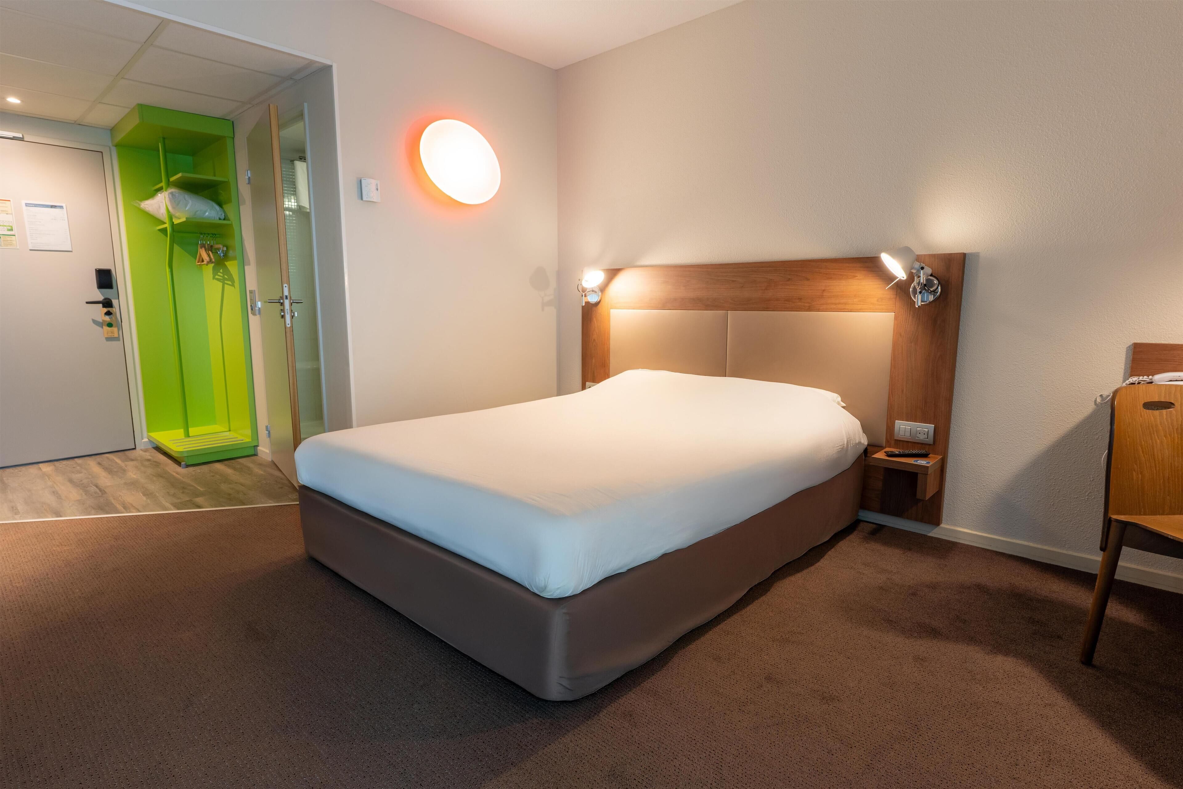 A hotel room with a large bed, a wooden headboard, bedside lamps, a nightstand, and a green door.