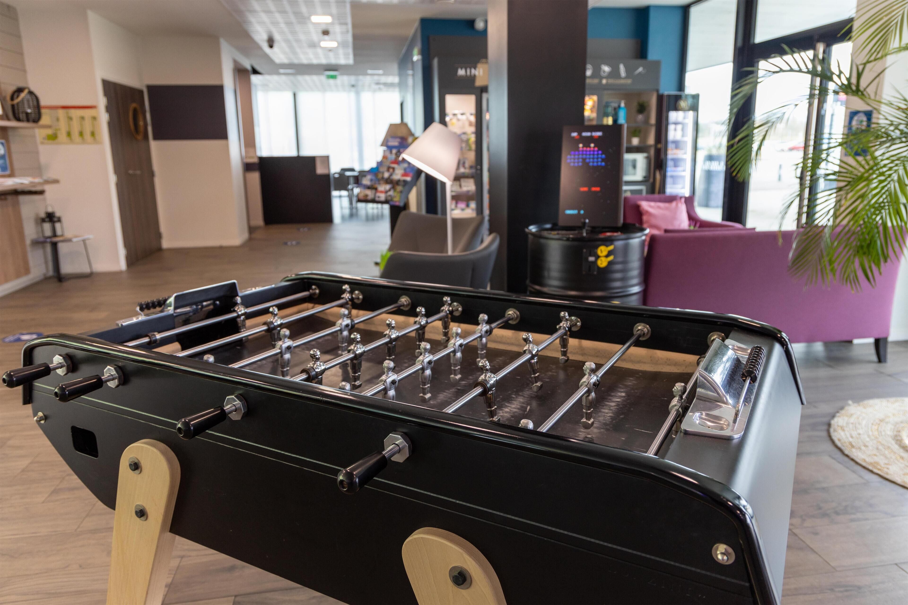 A foosball table in a modern lobby with a coffee machine, seating area, and a large window in the background.