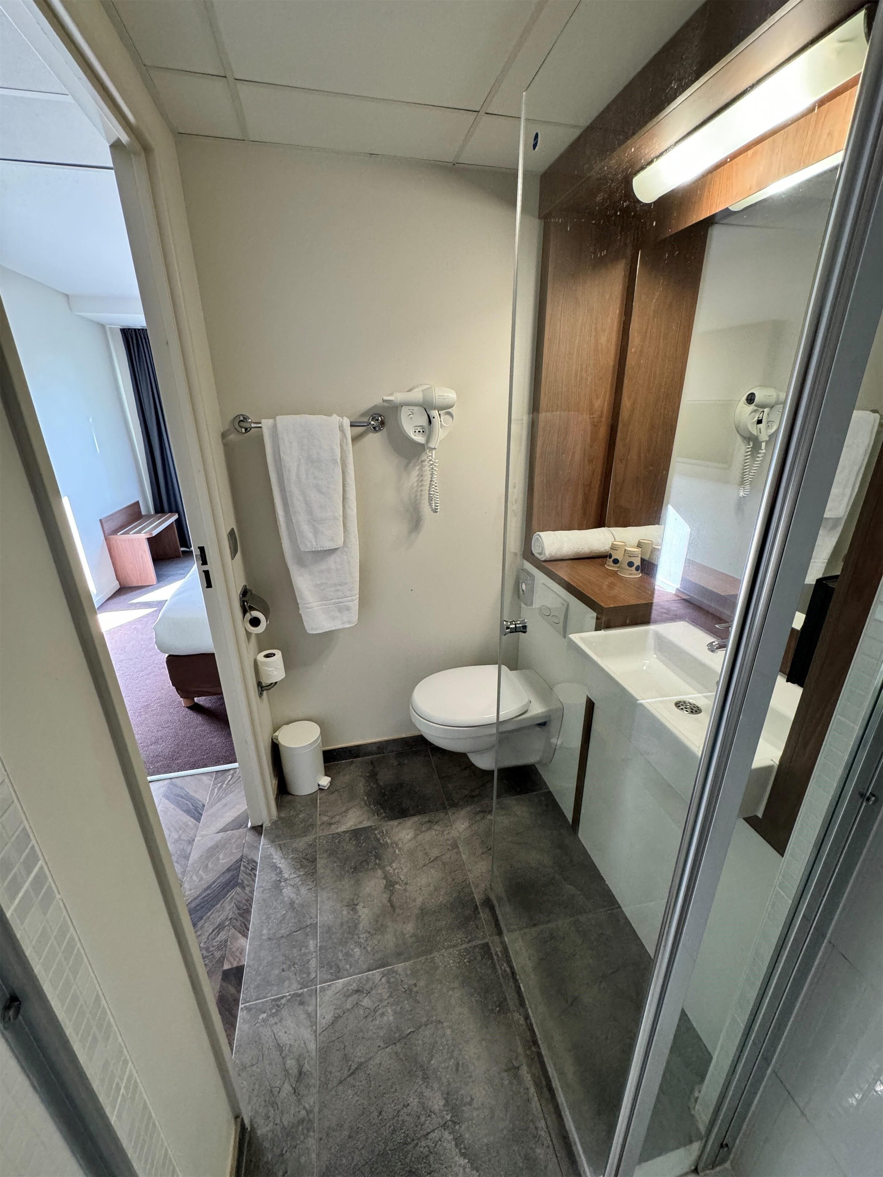 Un baño compacto con inodoro, cabina de ducha con mampara de vidrio, lavamanos y perchero para toallas.