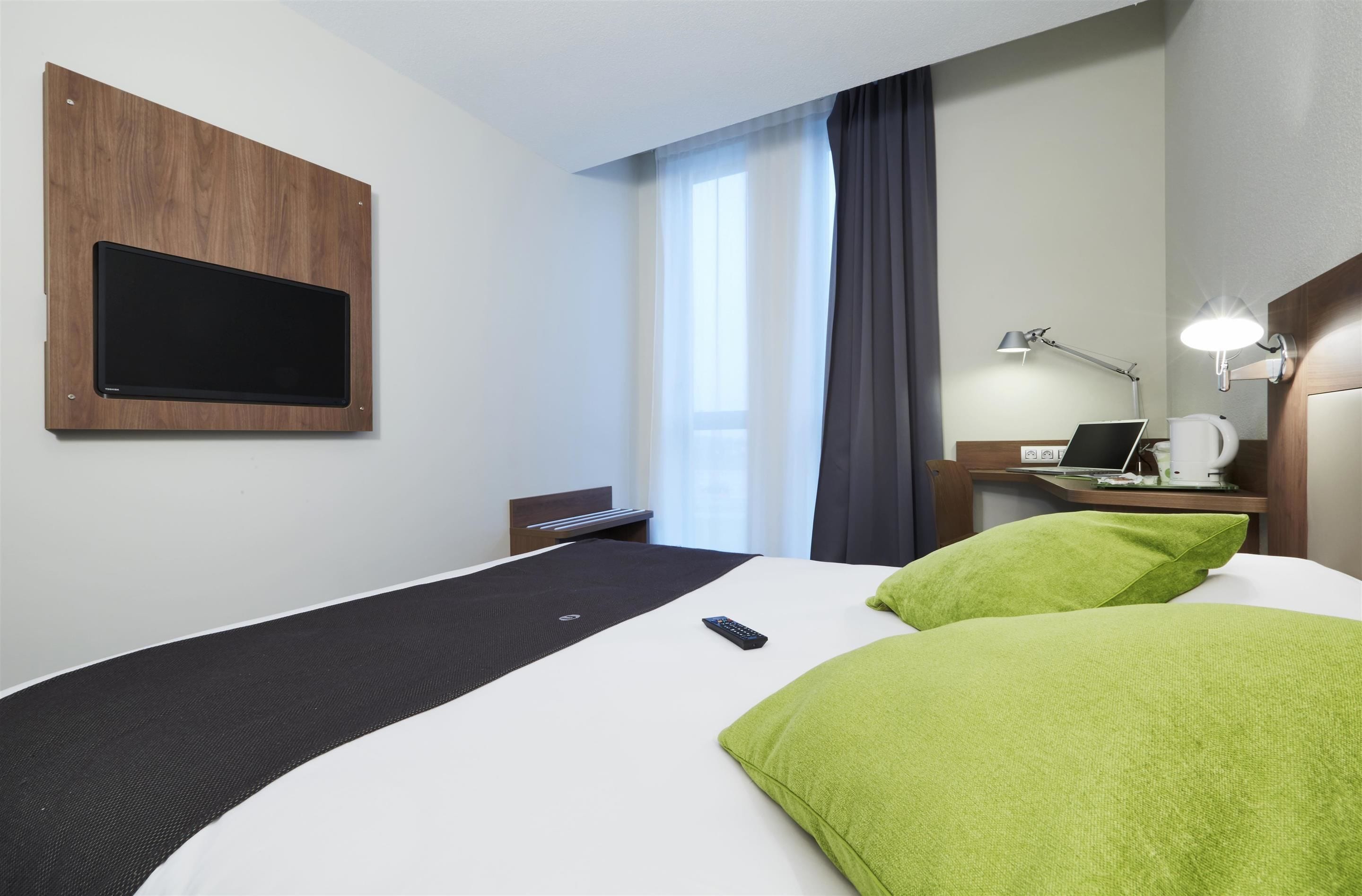 A hotel room with a bed, a desk with a laptop, a TV mounted on the wall, and a window with curtains.