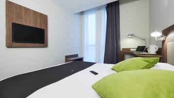 A hotel room with a bed, a desk with a laptop, a TV mounted on the wall, and a window with curtains.