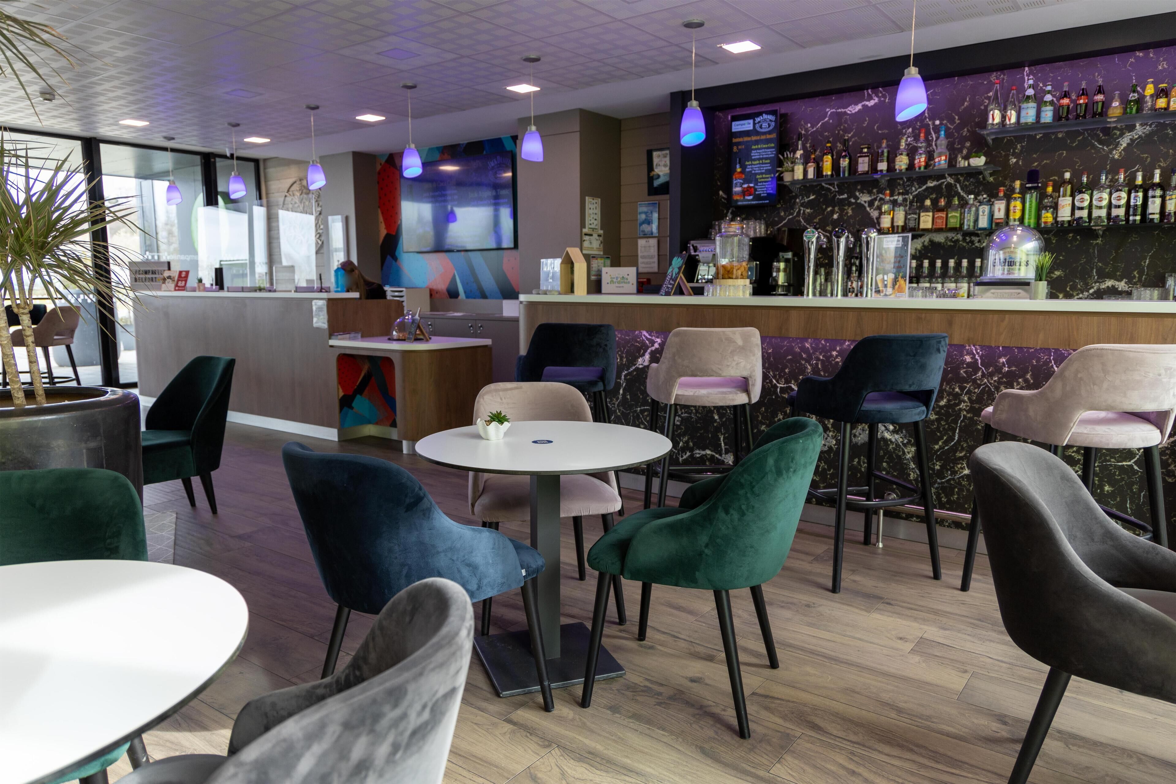 A modern bar area with a variety of seating options including round tables and chairs, a bar counter with a selection of bottles, and a large screen displaying content.