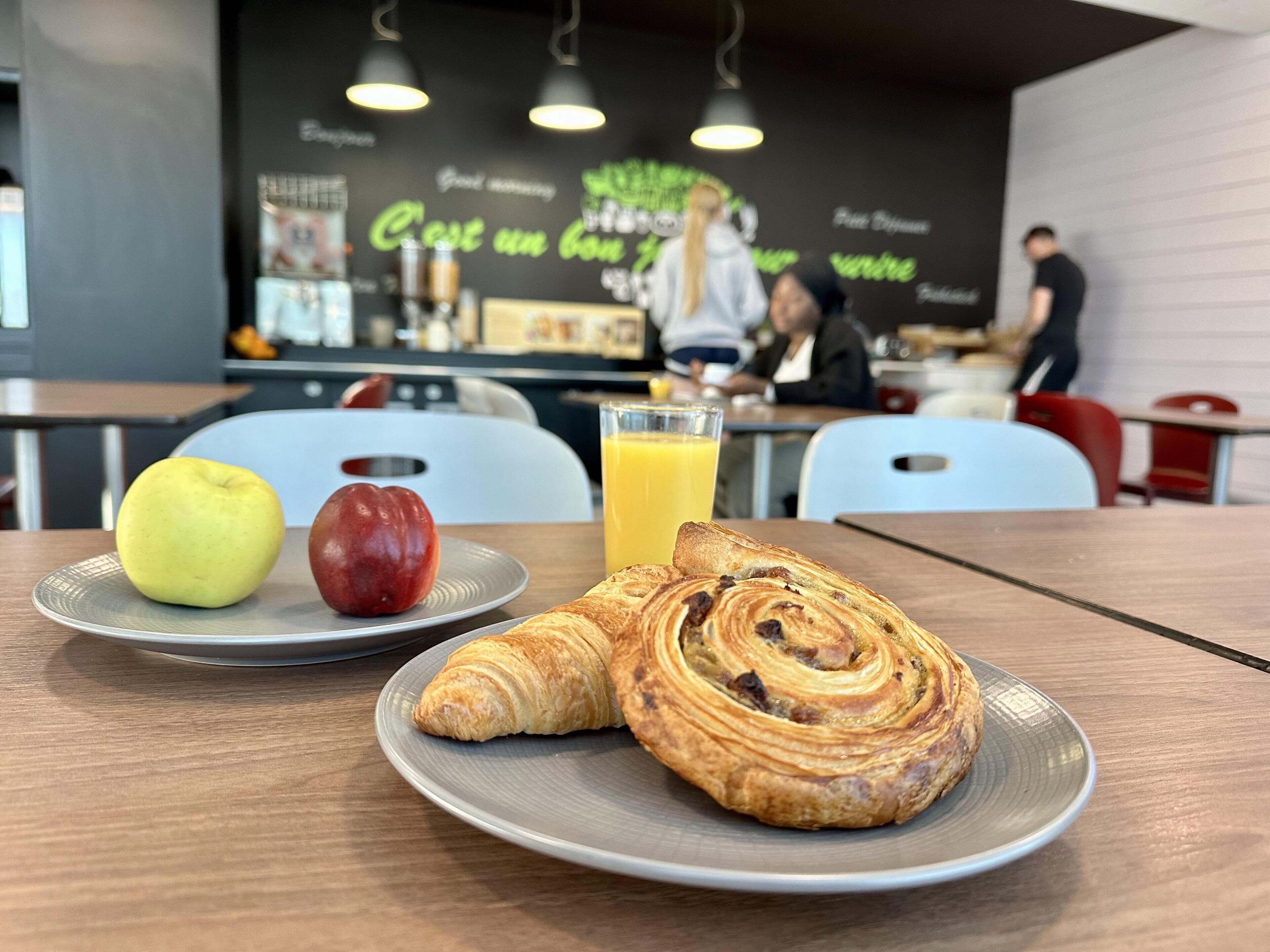 A breakfast setting with a croissant and two fruits on a plate, a glass of orange juice, and a modern interior with seating and a counter in the background.