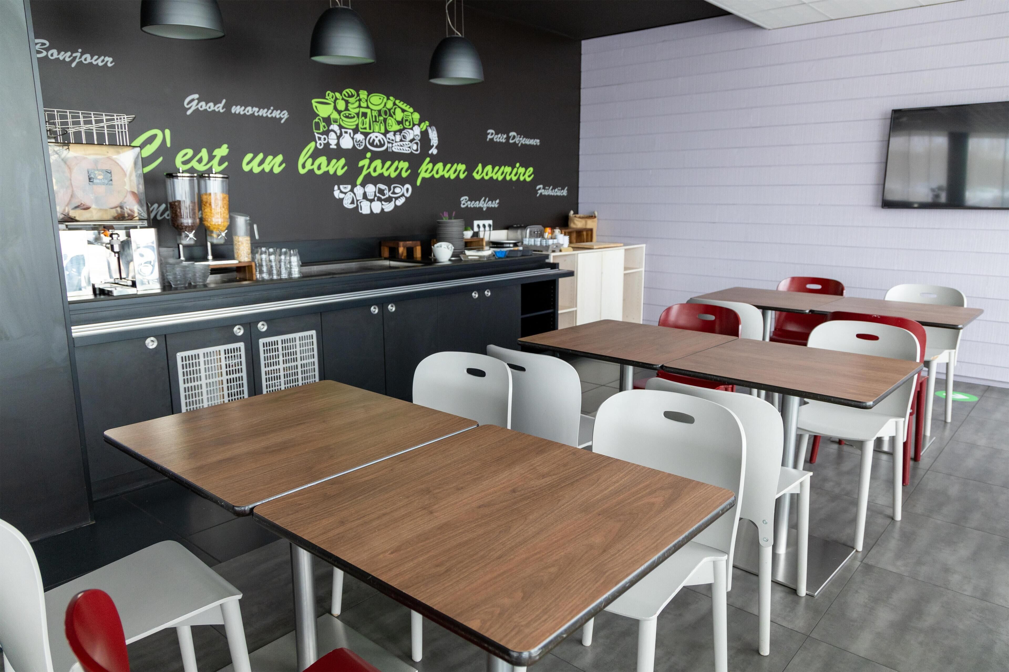 Un café moderne avec des tables en bois et des chaises blanches, un coin cuisine où sont préparés les plats du petit-déjeuner, et un mur orné de textes en français sur le thème du petit-déjeuner.