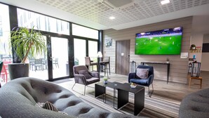 A modern living room with a large TV displaying a soccer game, comfortable seating, and a large window offering a view of the outside. - Campanile Belfort Montbéliard - Gare La Jonxion (Meroux-Moval)