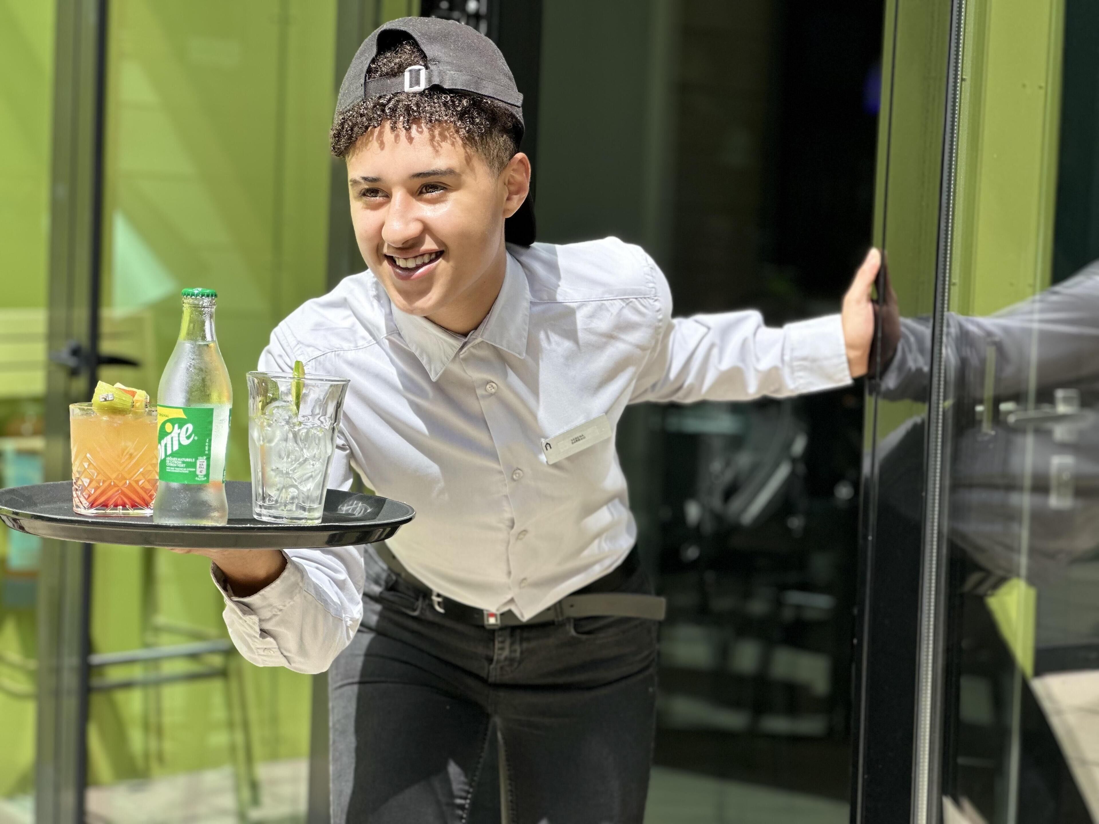 Een persoon draagt een dienblad met een frisdrankfles, een glas water en een drankje versierd met fruit.