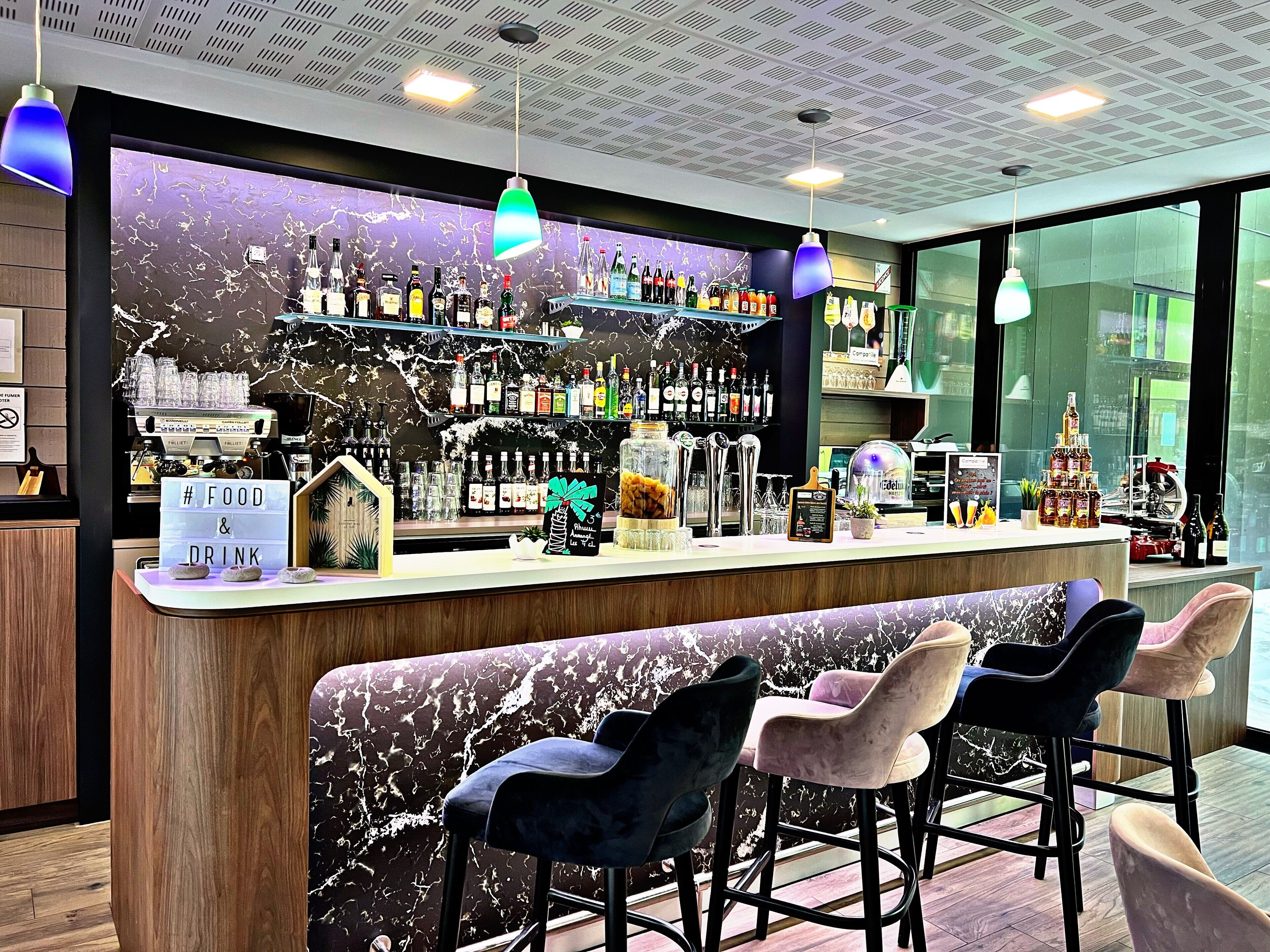 Un espace bar avec un mur recouvert de marbre, de nombreuses bouteilles d’alcool, une machine à café et des tabourets de bar.