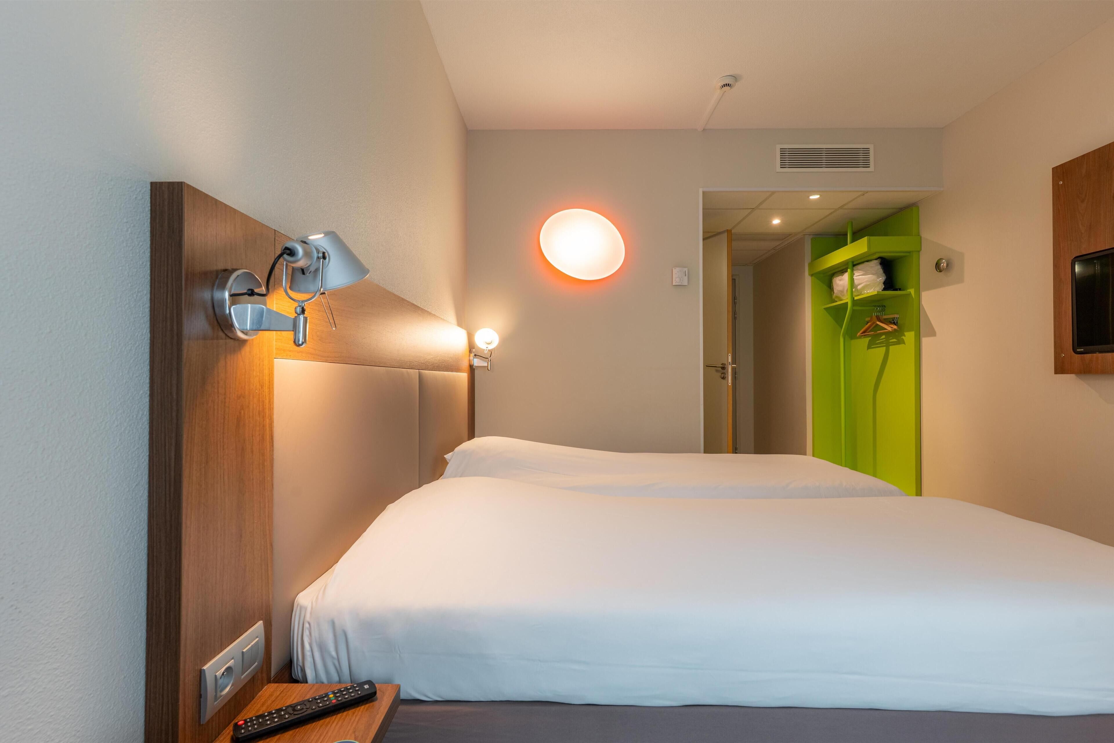 A hotel room with a bed, a desk with a lamp, a remote control, and a green storage unit.