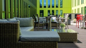 An outdoor seating area with wicker furniture, a glass-top table, and a view of a modern building with large windows. - Campanile Belfort Montbéliard - Gare La Jonxion (Meroux-Moval)