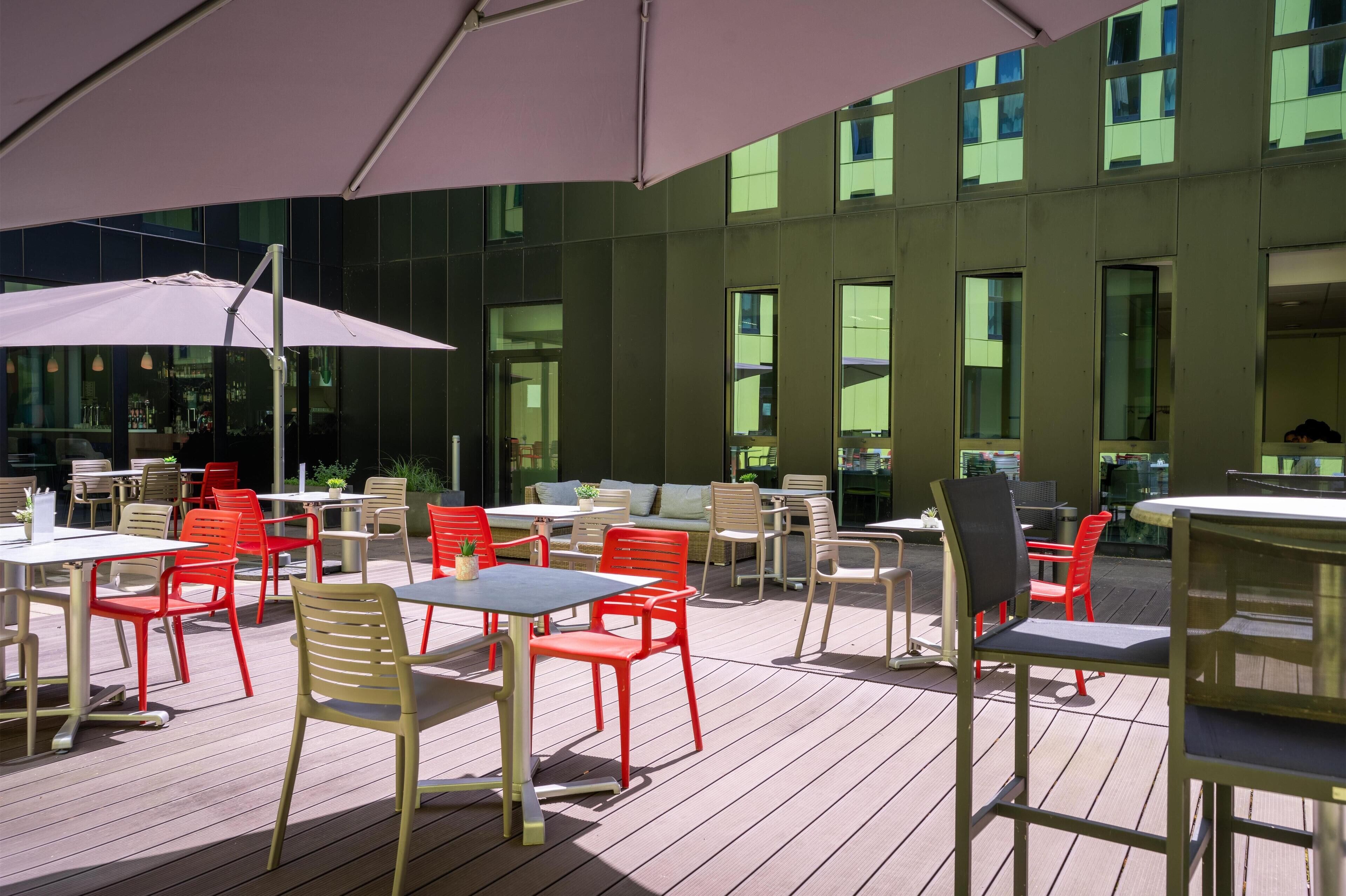 An outdoor patio area with tables and chairs, a large umbrella, and a modern building in the background.