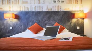 Habitación de hotel con una cama, dos almohadas naranjas, una computadora portátil y una revista sobre la cama.