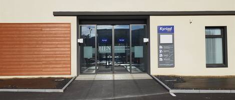 A hotel entrance with a glass door, a wooden wall on the left, and a sign for 'Kyriad Hotel' on the right.