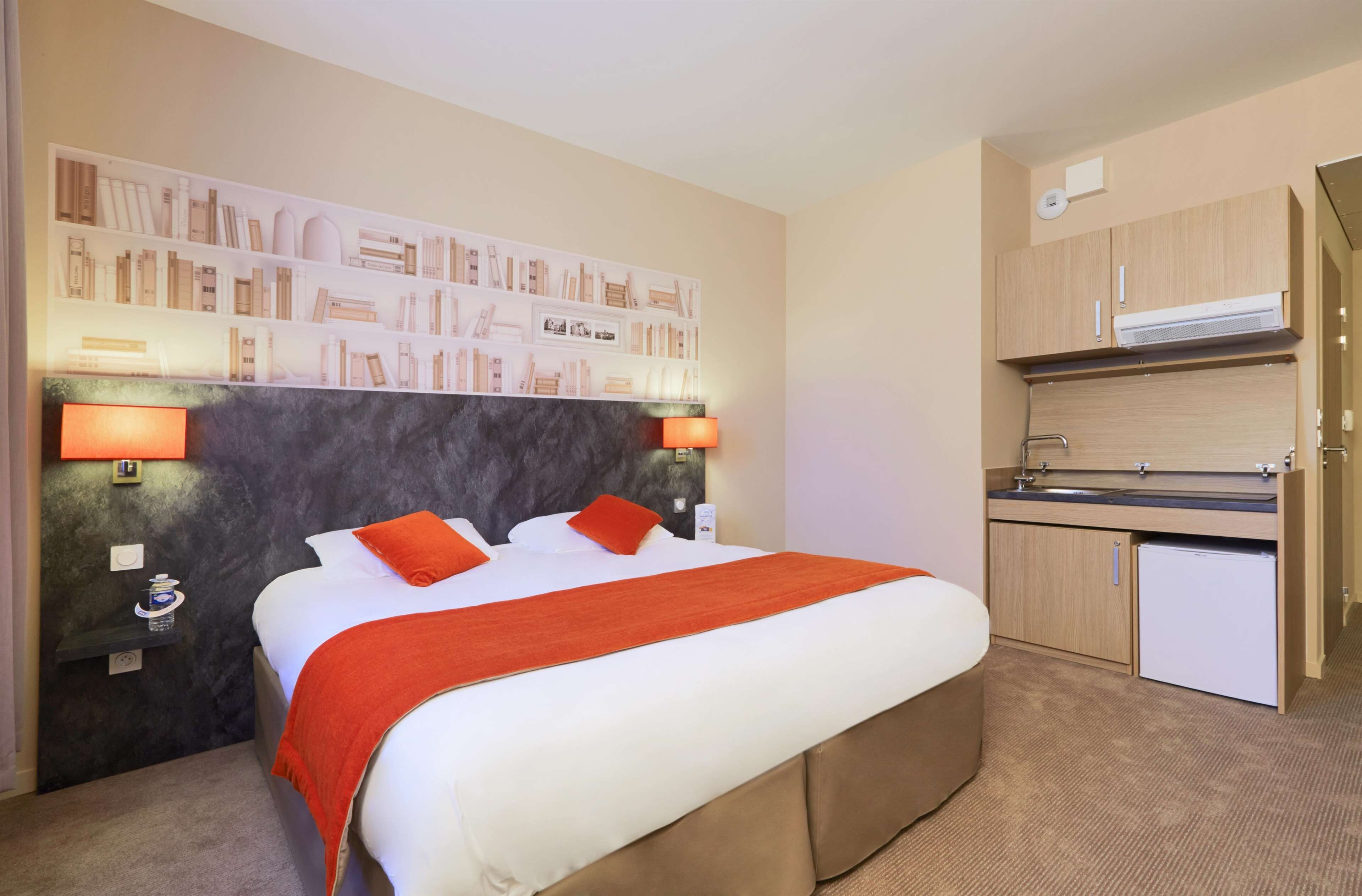 A hotel room with a bed, a small kitchenette, and a wall with a bookshelf.