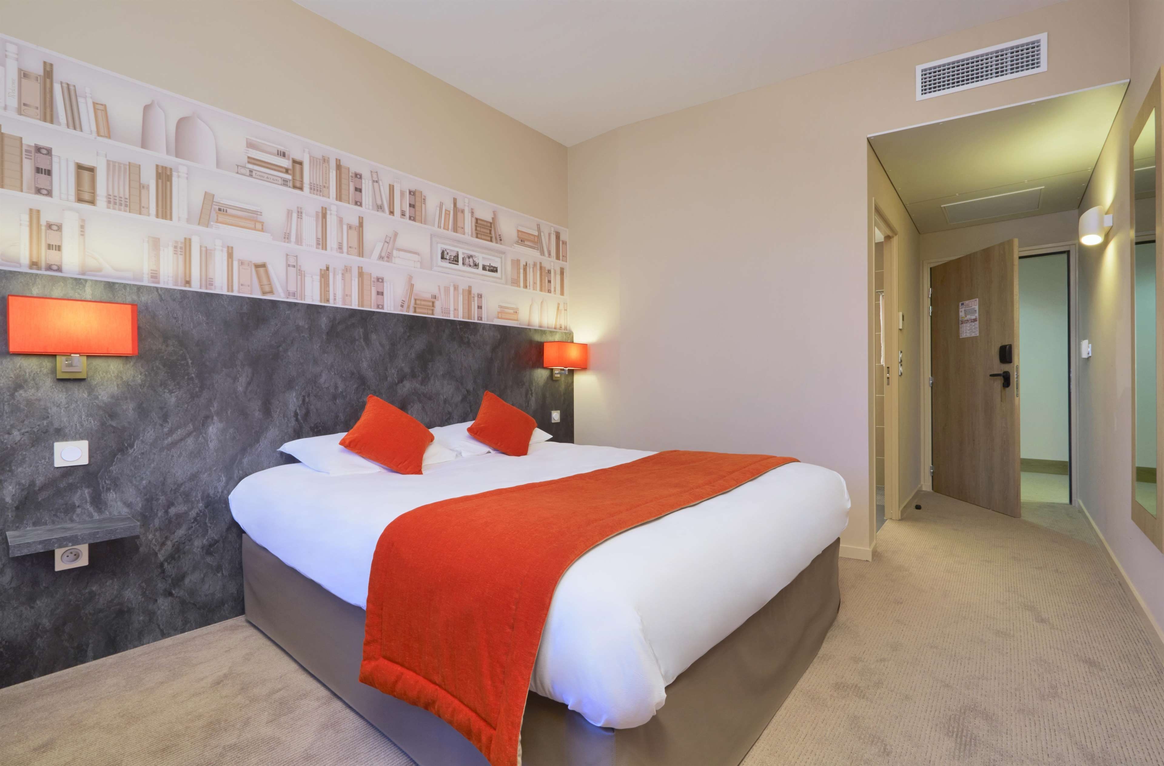 Una habitación de hotel con una cama grande, dos lámparas de noche, una estantería y una puerta que da a otra habitación.