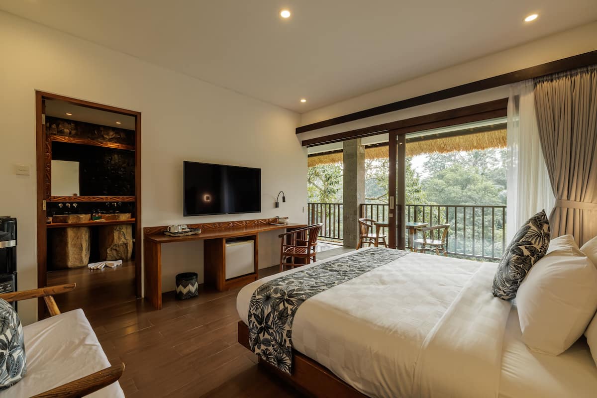 Ein Schlafzimmer mit einem großen Bett, einem Fernseher, einem Schreibtisch und einem Balkon mit Blick auf Bäume.