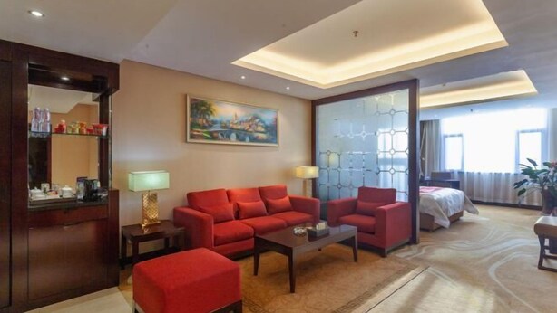 Habitación de hotel con sofá rojo, una mesita de centro, escritorio con lámpara, un cuadro en la pared y una ventana grande con vistas.