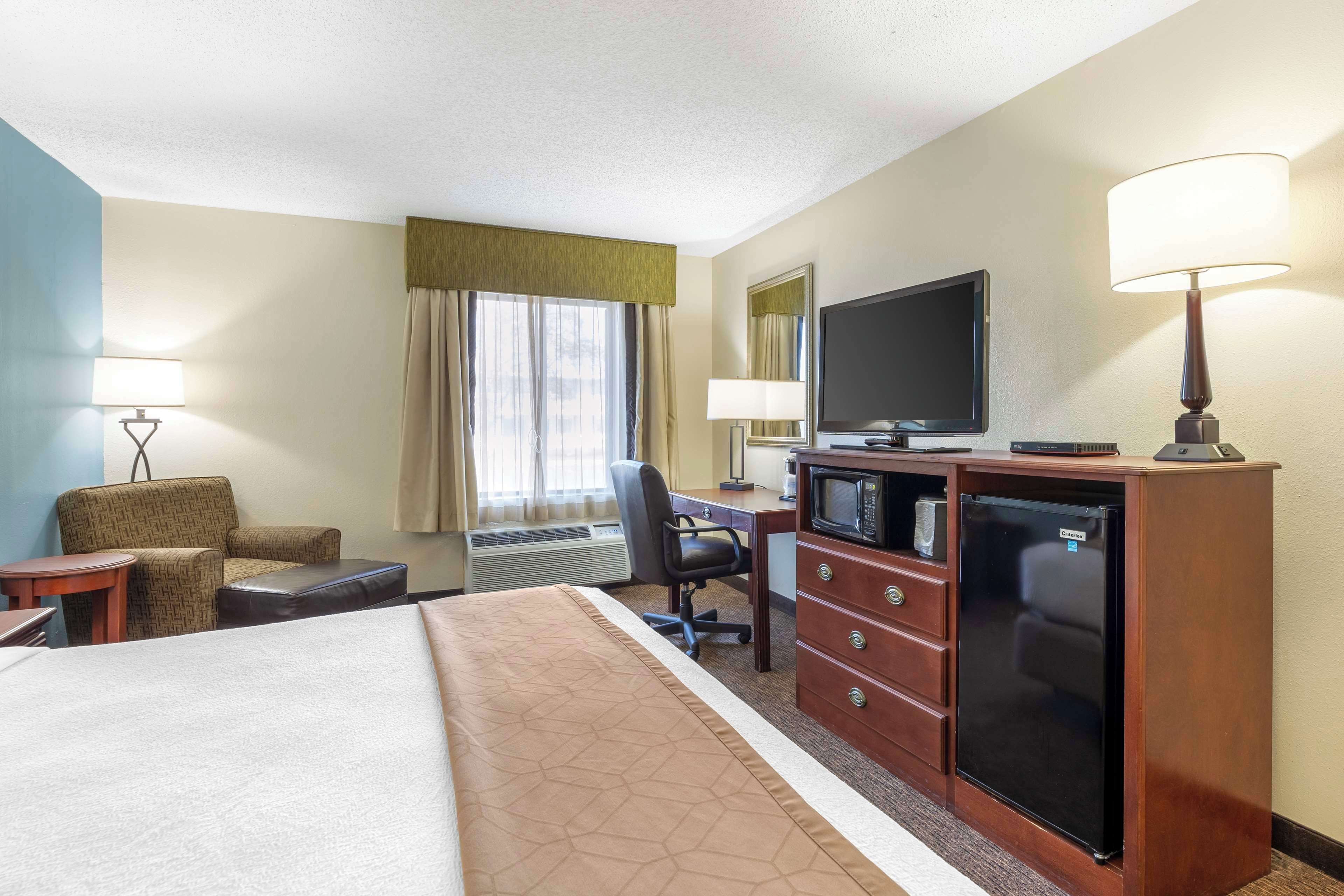 A hotel room with a bed, a desk with a computer, a television, a refrigerator, a chair, and a lamp.