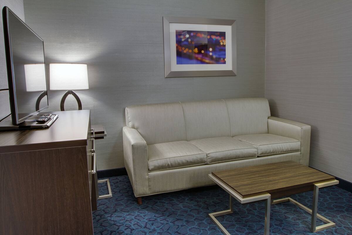 A hotel room with a sofa, a small table, a desk with a lamp, and a framed picture on the wall.