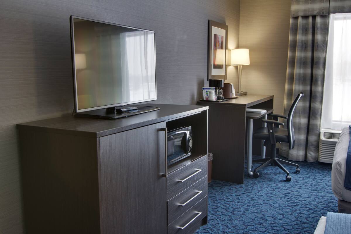 A hotel room with a flat-screen TV on a cabinet, a desk with a lamp, a chair, and a bed with a blue carpet.