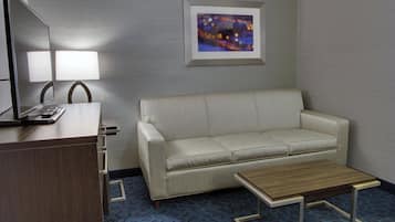 A hotel room with a sofa, a small table, a desk with a lamp, and a framed picture on the wall.