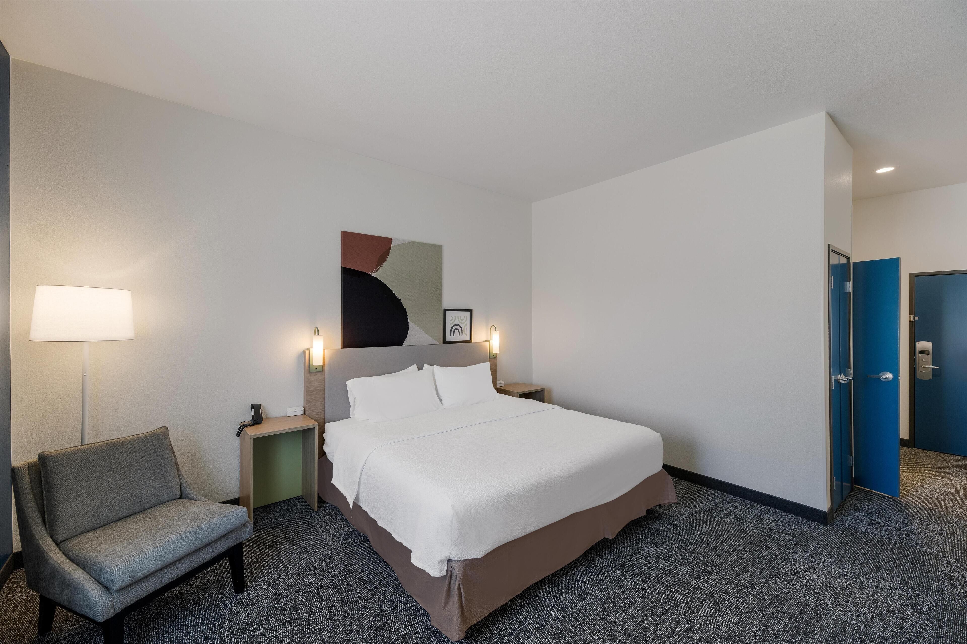 A hotel room with a bed, a chair, a nightstand, a lamp, and a door.