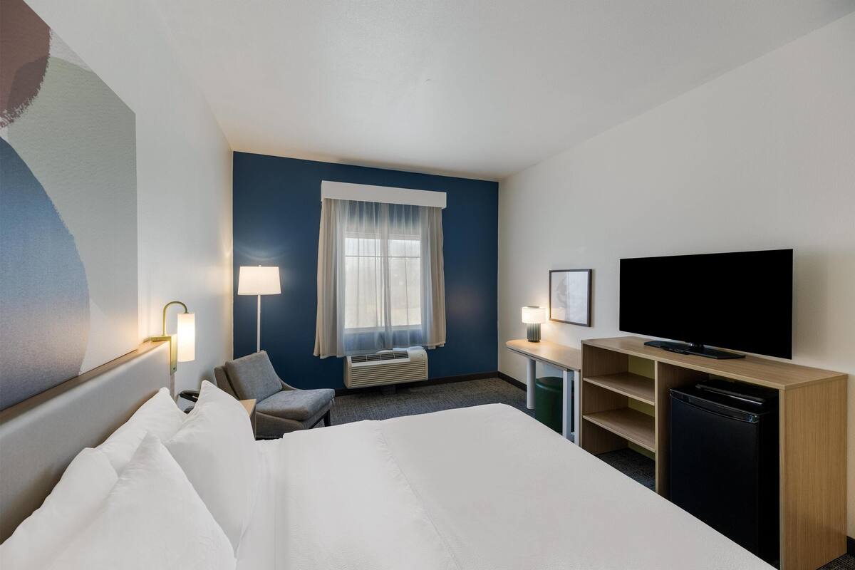 A hotel room with a large bed, a desk with a flat-screen TV, a chair, a lamp, and a window with curtains.