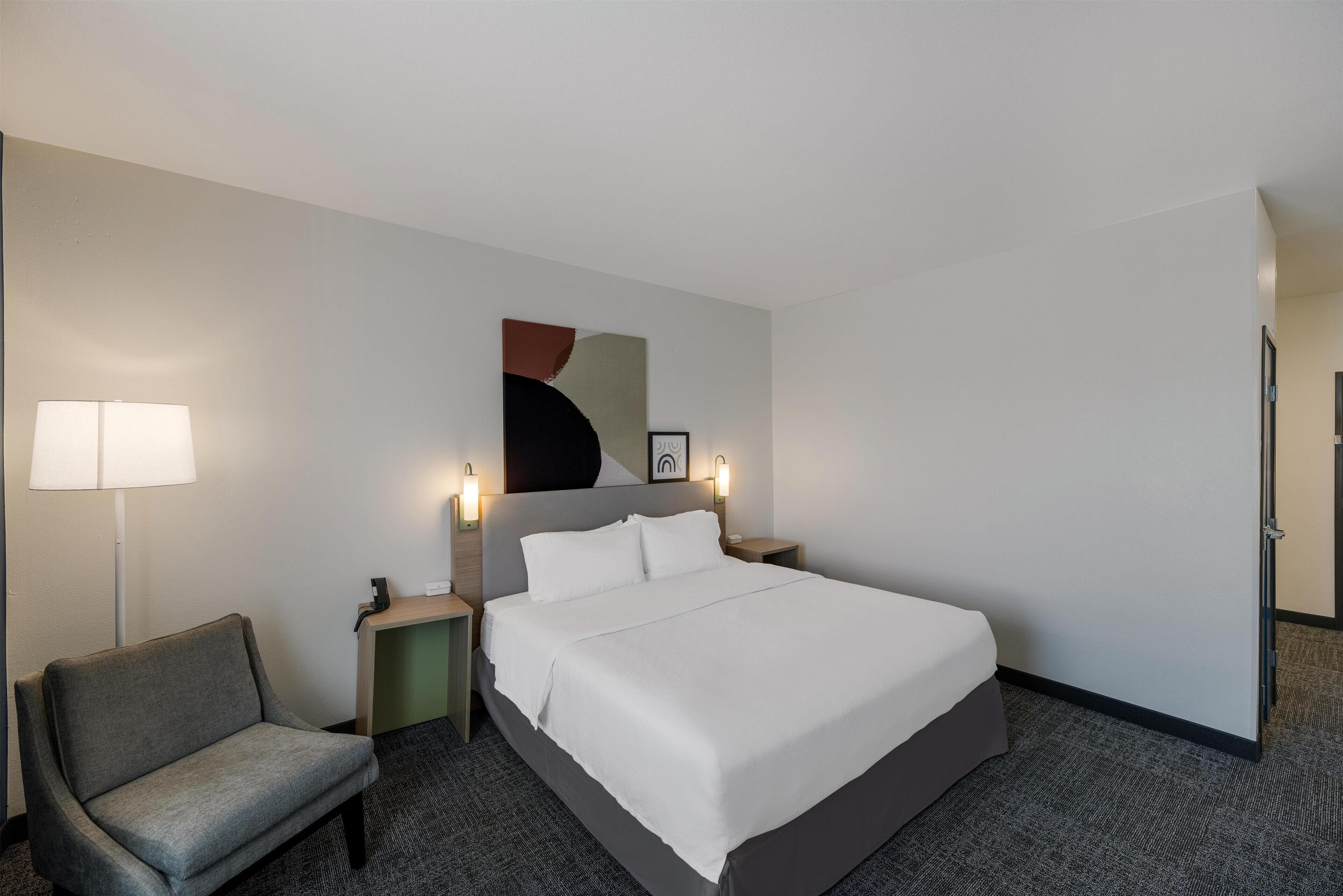 A hotel room with a bed, a chair, a desk, and a lamp.