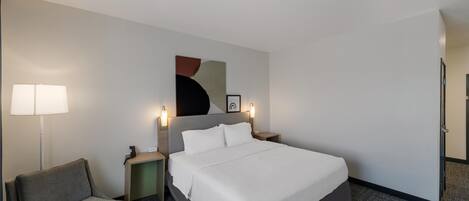 A hotel room with a bed, a chair, a desk, and a lamp.