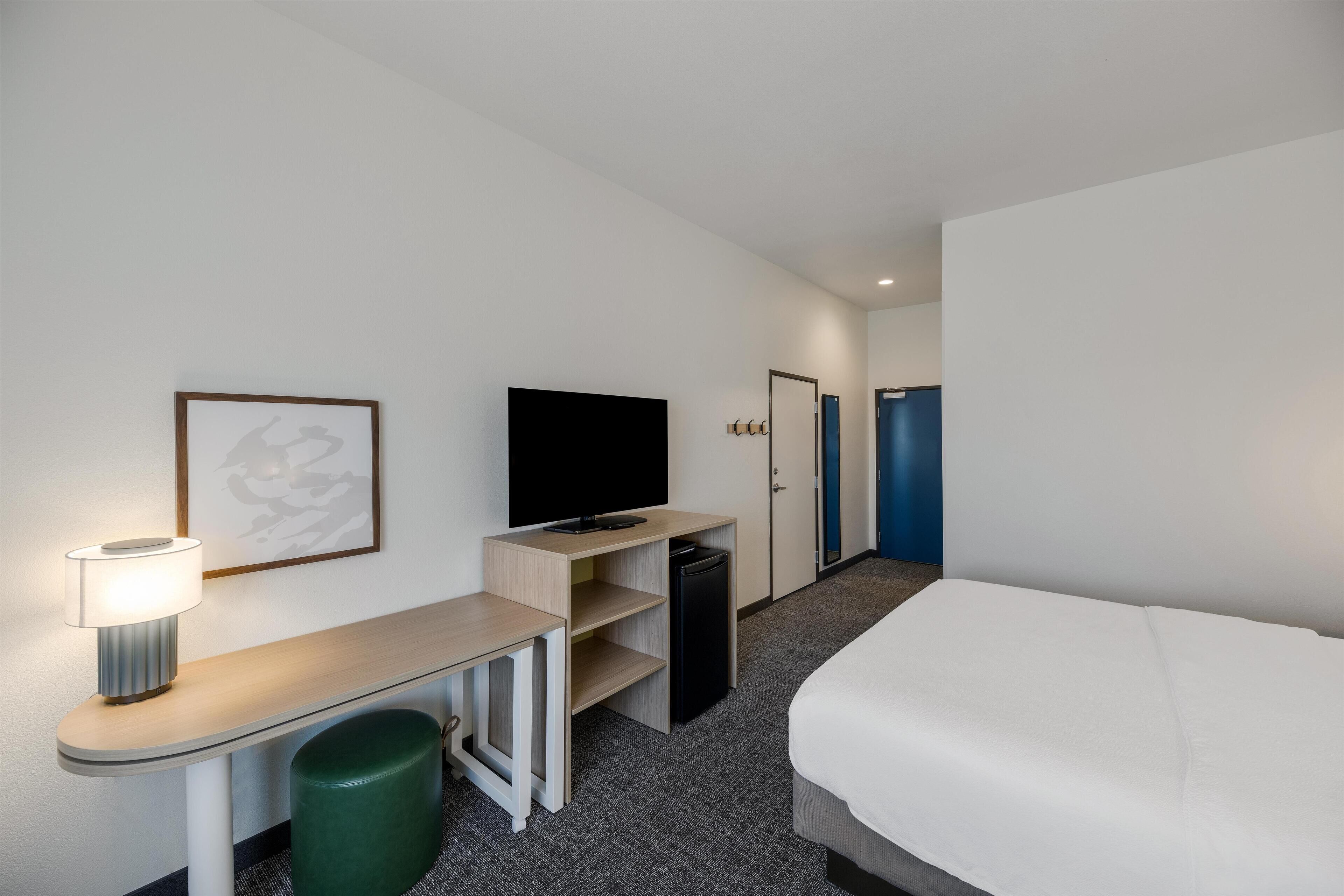 Una habitación de hotel con una cama, un escritorio con lámpara, televisor, cesto de basura y una puerta que da a otra habitación.