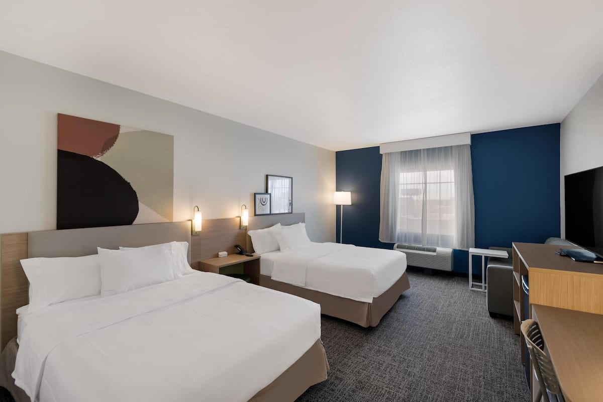 A hotel room with two beds, a desk, a chair, a lamp, and a large abstract painting on the wall.