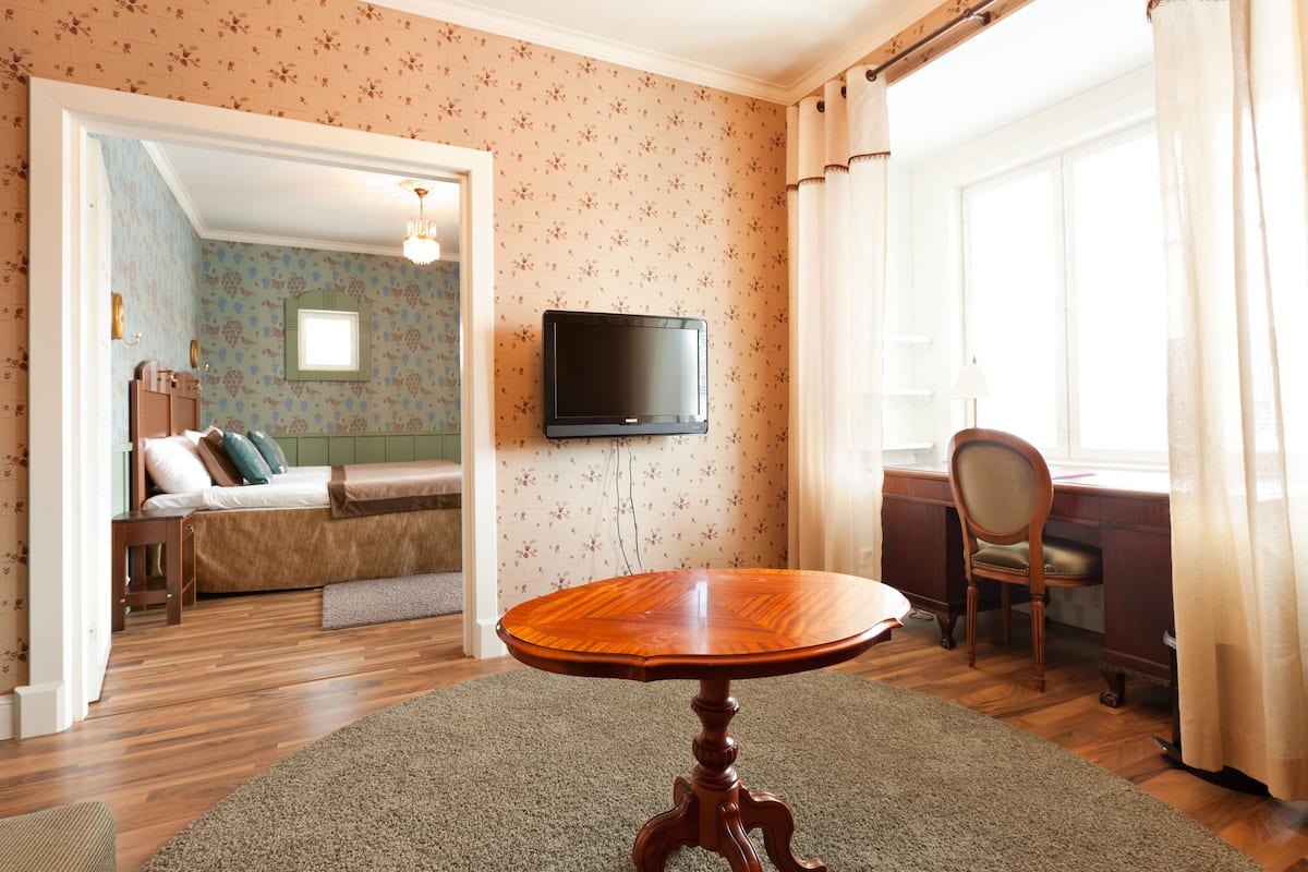 A hotel room with a wooden floor, a round wooden table, a bed with a green headboard, a TV mounted on the wall, and a window with curtains.
