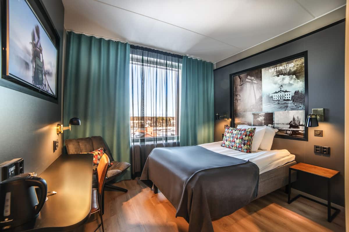 A hotel room with a bed, a desk, a chair, a small table, a window with curtains, and a wall-mounted picture.