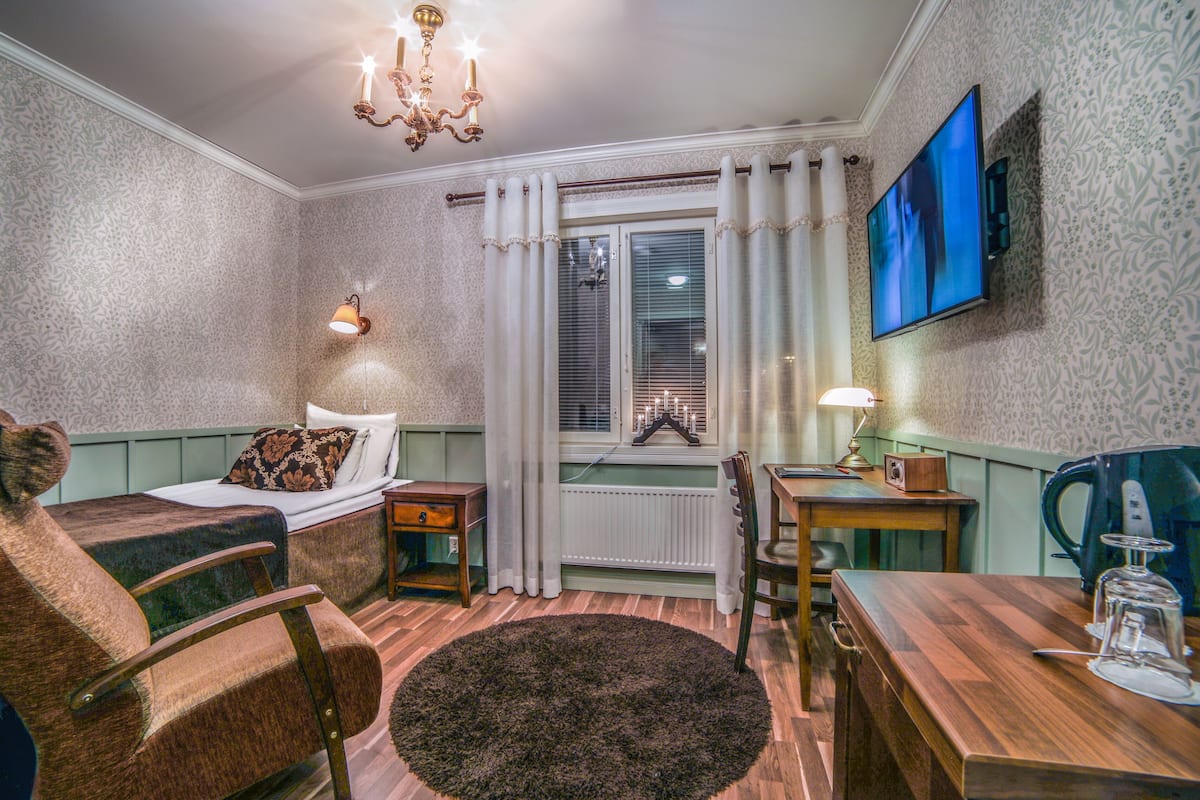 A cozy room with a bed, a chair, a table, a TV, and a window with curtains.