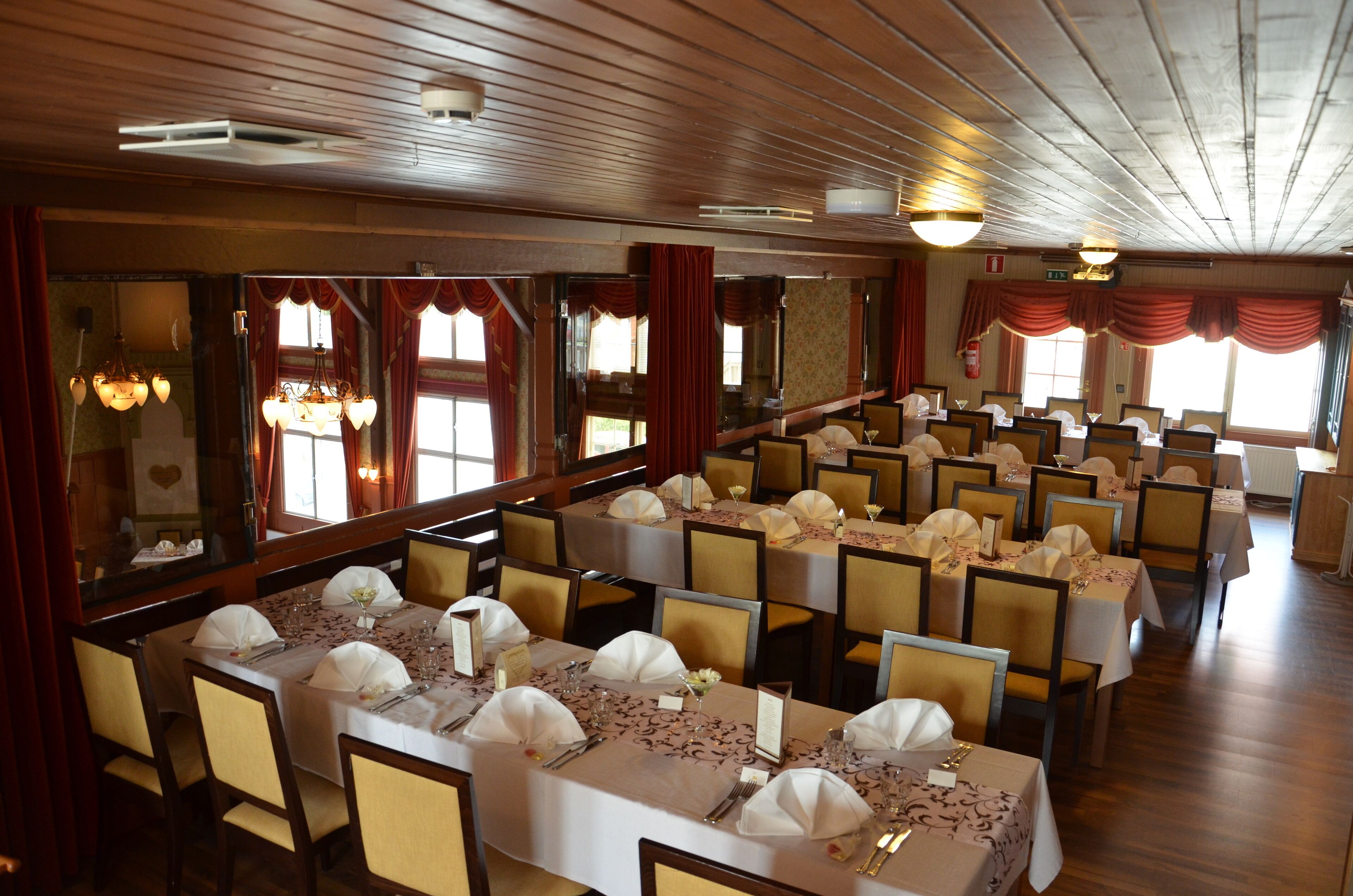 A banquet hall with multiple tables set for dining, wooden paneling on the ceiling, and large windows with curtains.