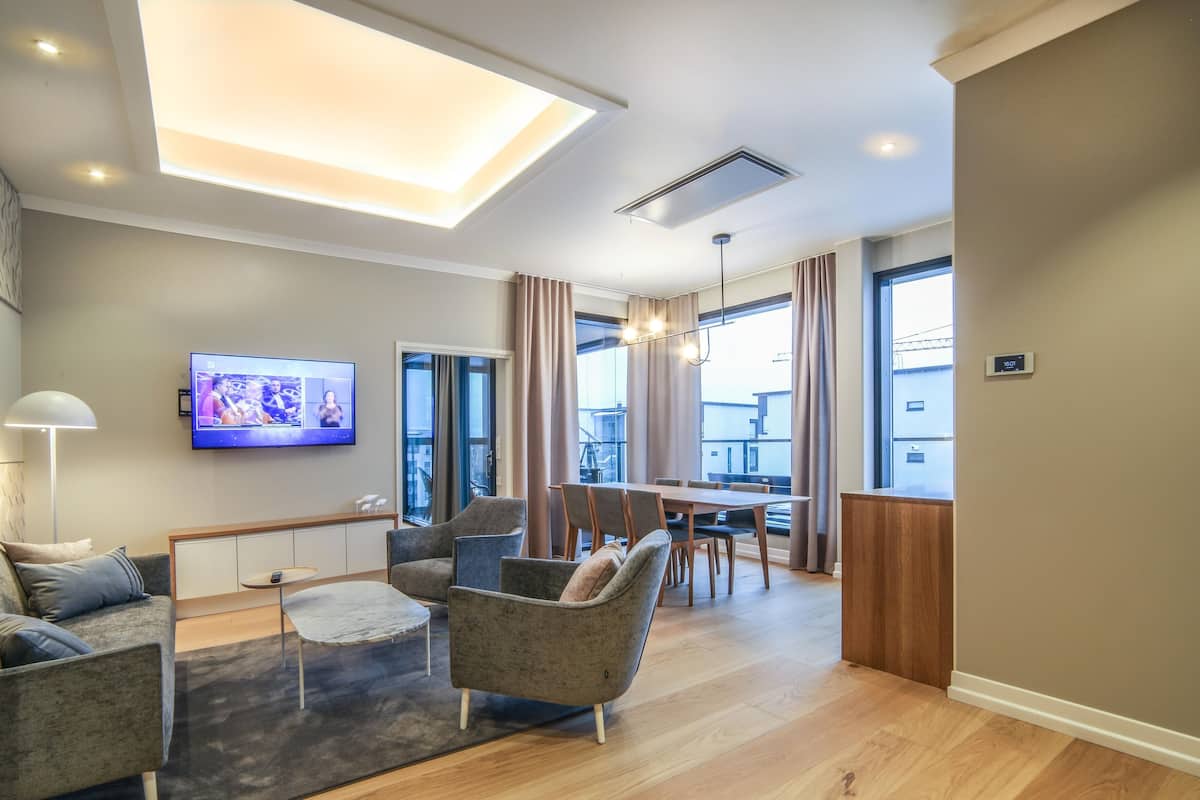 A modern living room with a flat-screen TV, comfortable seating, a dining area, and large windows.