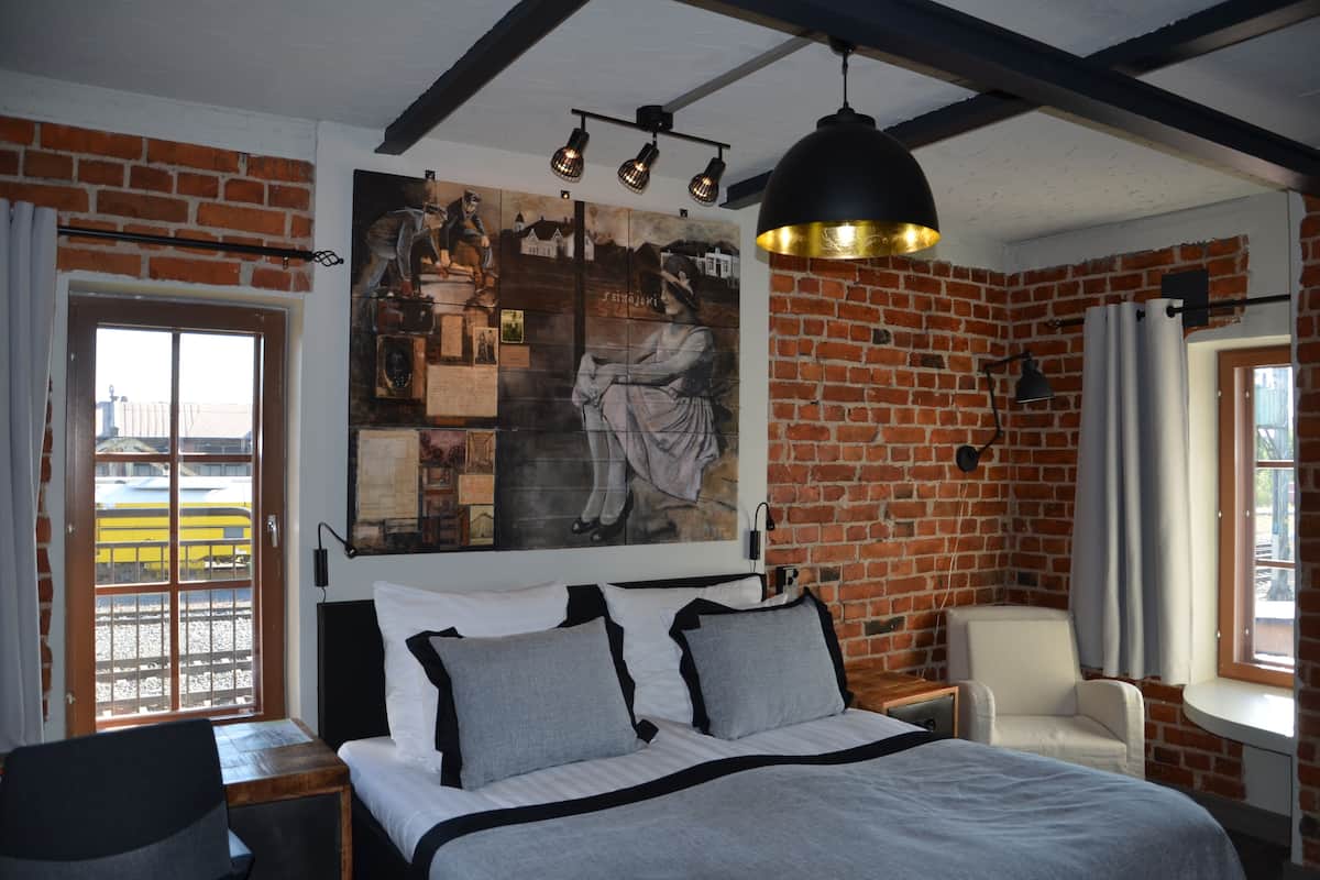 A bedroom with a brick wall, a large bed with grey bedding, a white chair, a desk, and a large painting.