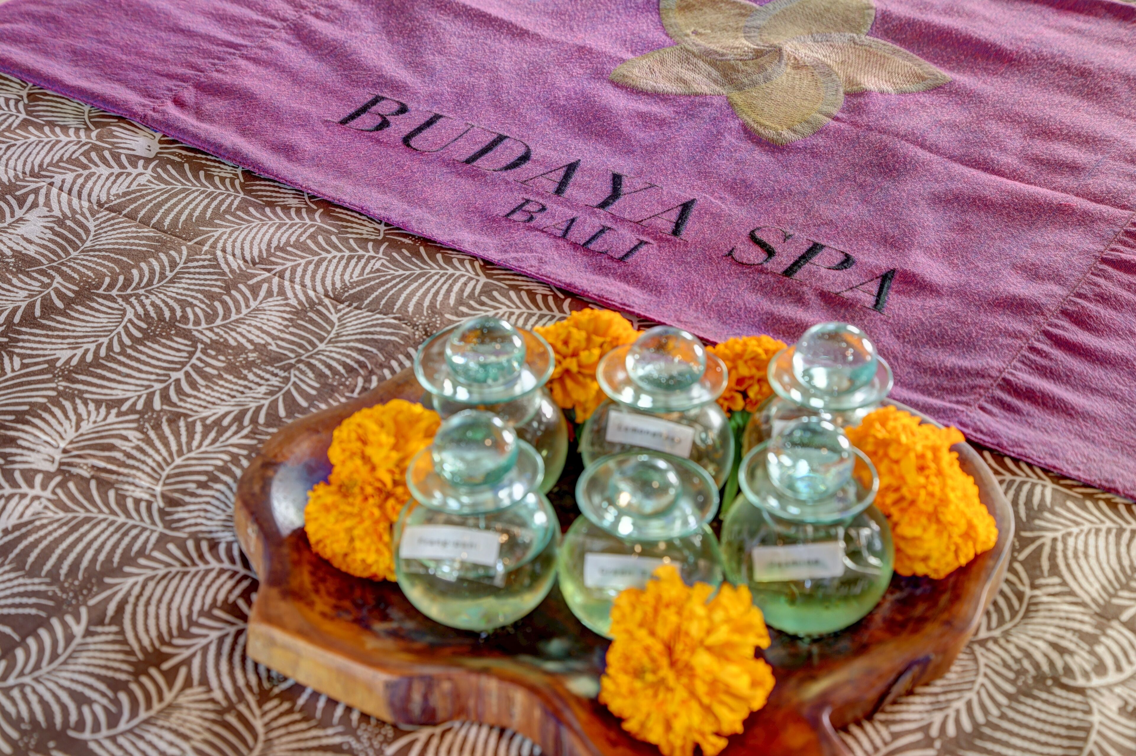 A tray of glass containers with labels, surrounded by marigold flowers, on a patterned fabric with a purple towel in the background.