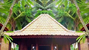 A wooden structure with a tiled roof, surrounded by lush greenery and palm trees. - Junjungan Suite Villa (Ubud)