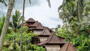 A traditional thatched-roof house with a wooden balcony, surrounded by lush greenery and palm trees. - Junjungan Suite Villa (Ubud)